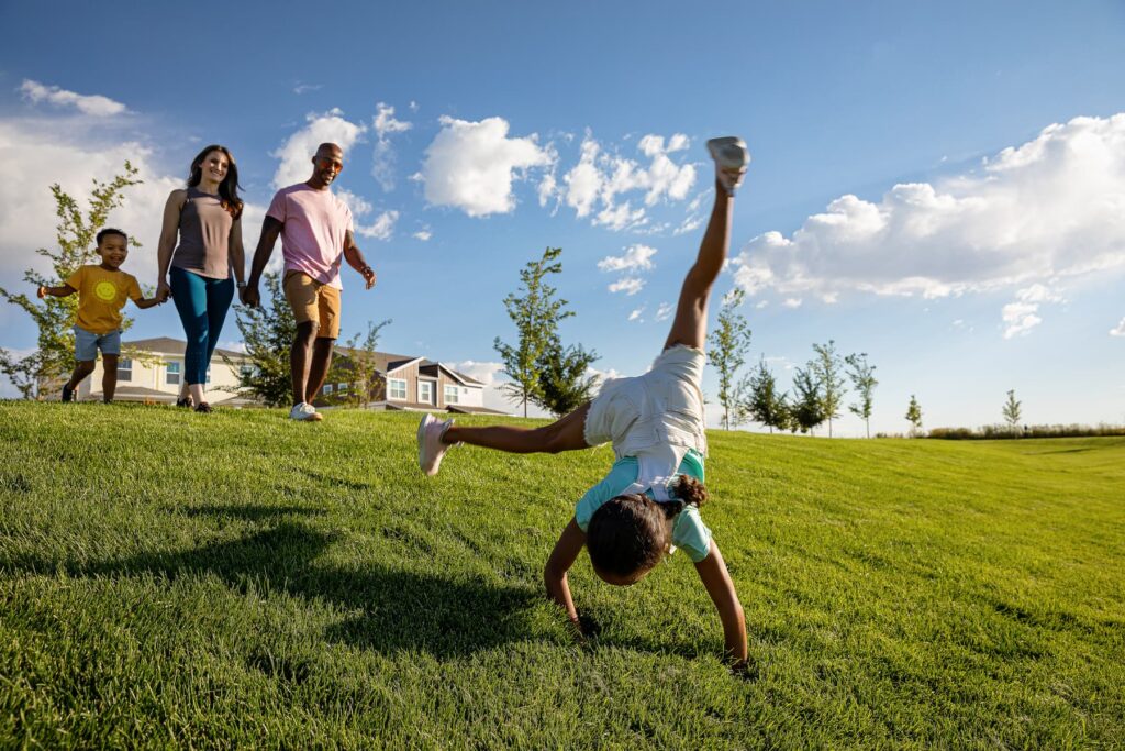 A family of four enjoys a sunny day on the grass as a young girl does a cartwheel, framed by trees and uptown homes for sale in the background—capturing the vibrant spirit of new homes in Aurora, CO.