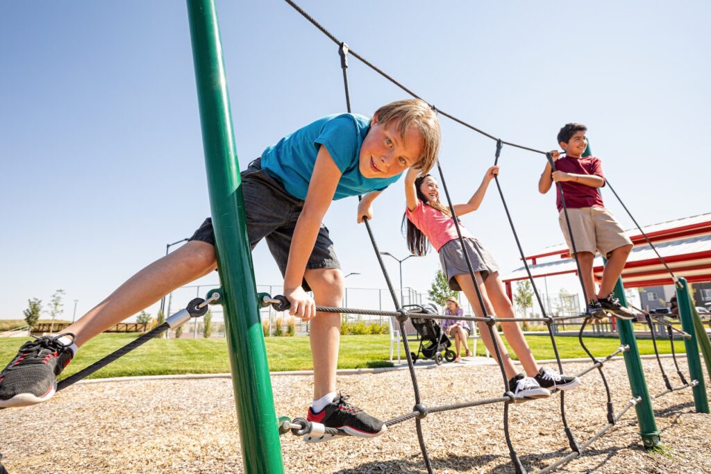 Three children climb on a rope structure at a playground on a sunny day, with grass and park equipment visible in the background—an inviting scene near Horizon Uptown Aurora CO, one of the area’s vibrant new home communities.