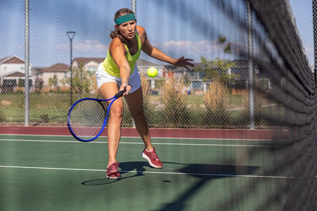 A woman in athletic wear stretches to hit a tennis ball with a racket on an outdoor court, seen through the net, capturing the active lifestyle at Horizon Uptown aurora co.
