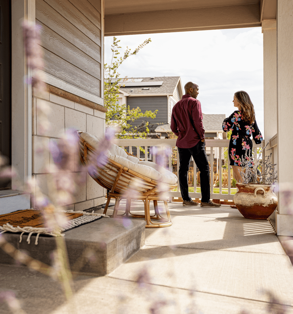 A man and woman stand talking on a front porch next to a wicker chair and large potted plant, with new construction homes in Aurora CO, like those in Horizon Uptown, visible in the background.