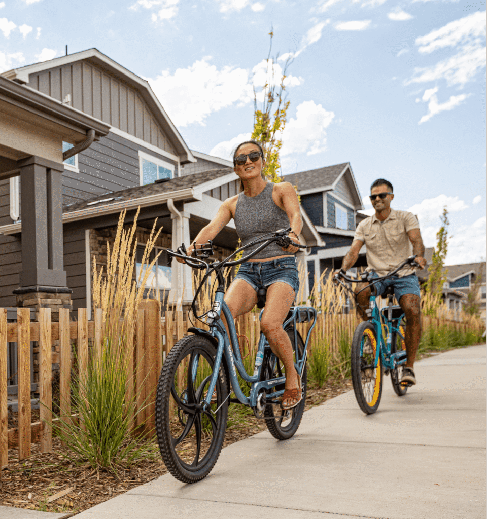 Two adults ride bicycles on a sunny day through a suburban neighborhood, passing houses and a wooden fence—capturing the inviting feel of new construction homes in Aurora CO.