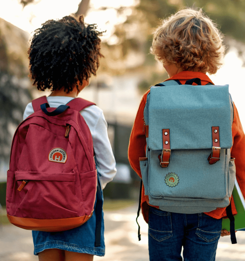 Two children with backpacks walk side by side outdoors in Horizon Uptown Aurora CO. One wears a red backpack, the other blue-gray, as they head toward new home communities, walking away from the camera.