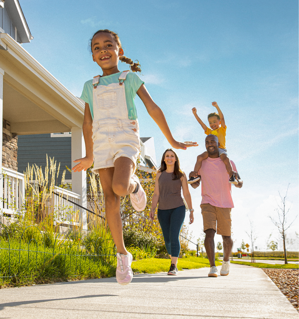 A young girl runs on a sidewalk in front of a house at Horizon Uptown Aurora, while a man carries a child on his shoulders and a woman walks beside them on a sunny day.
