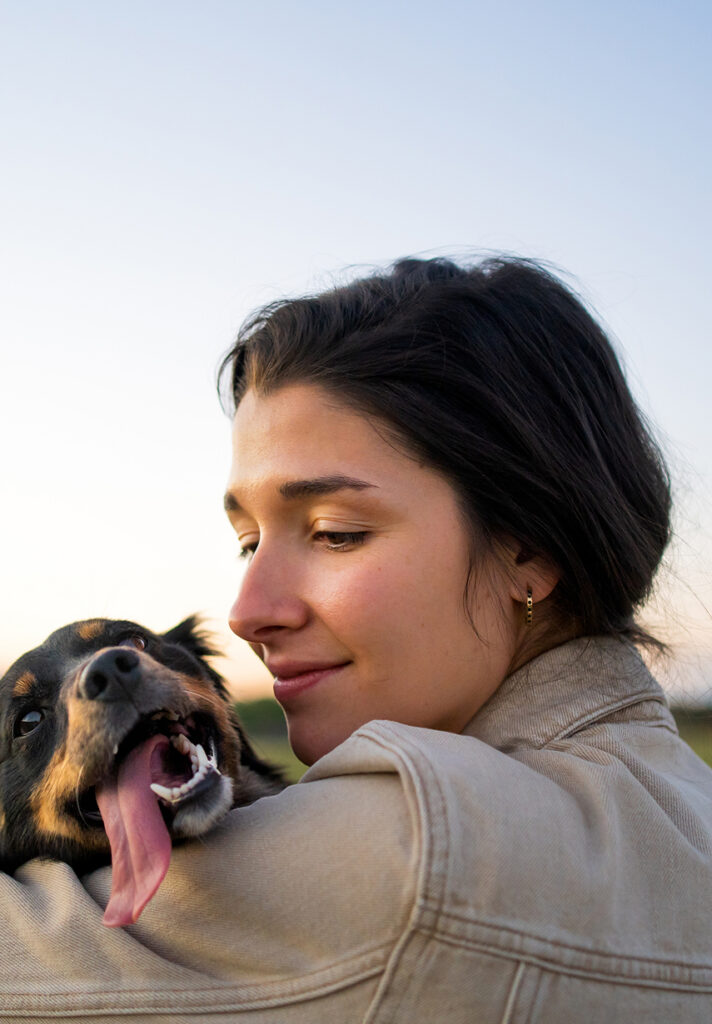 A woman with dark hair in a beige jacket hugs a happy dog with its tongue out, celebrating near new homes in Aurora, CO. The background shows a clear sky at dusk.
