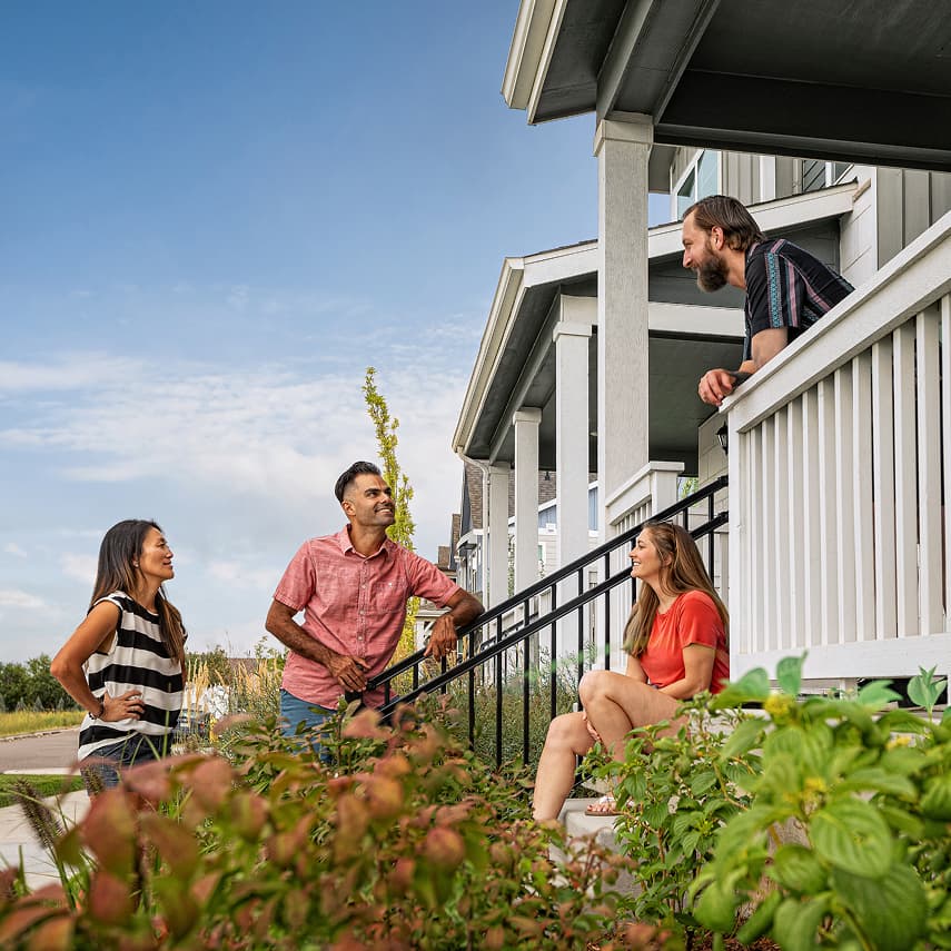 Four adults have a conversation outside on the steps and porch of a house in one of the beautiful aurora co new home communities, enjoying a sunny day in their suburban neighborhood.