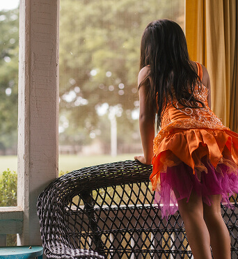A young girl in an orange and purple dress stands on a wicker chair, looking out a window toward trees outside, dreaming of adventures in Horizon Uptown Aurora CO or other new homes in Aurora CO.