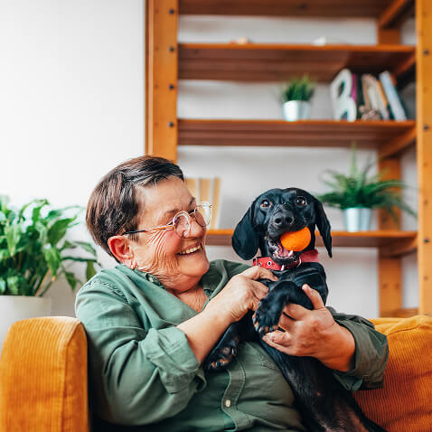 An older woman sits on a sofa, smiling as she holds a black dog with a ball in its mouth. Shelves and plants are visible in the background, creating a cozy scene that feels right at home in Horizon Uptown, Aurora.
