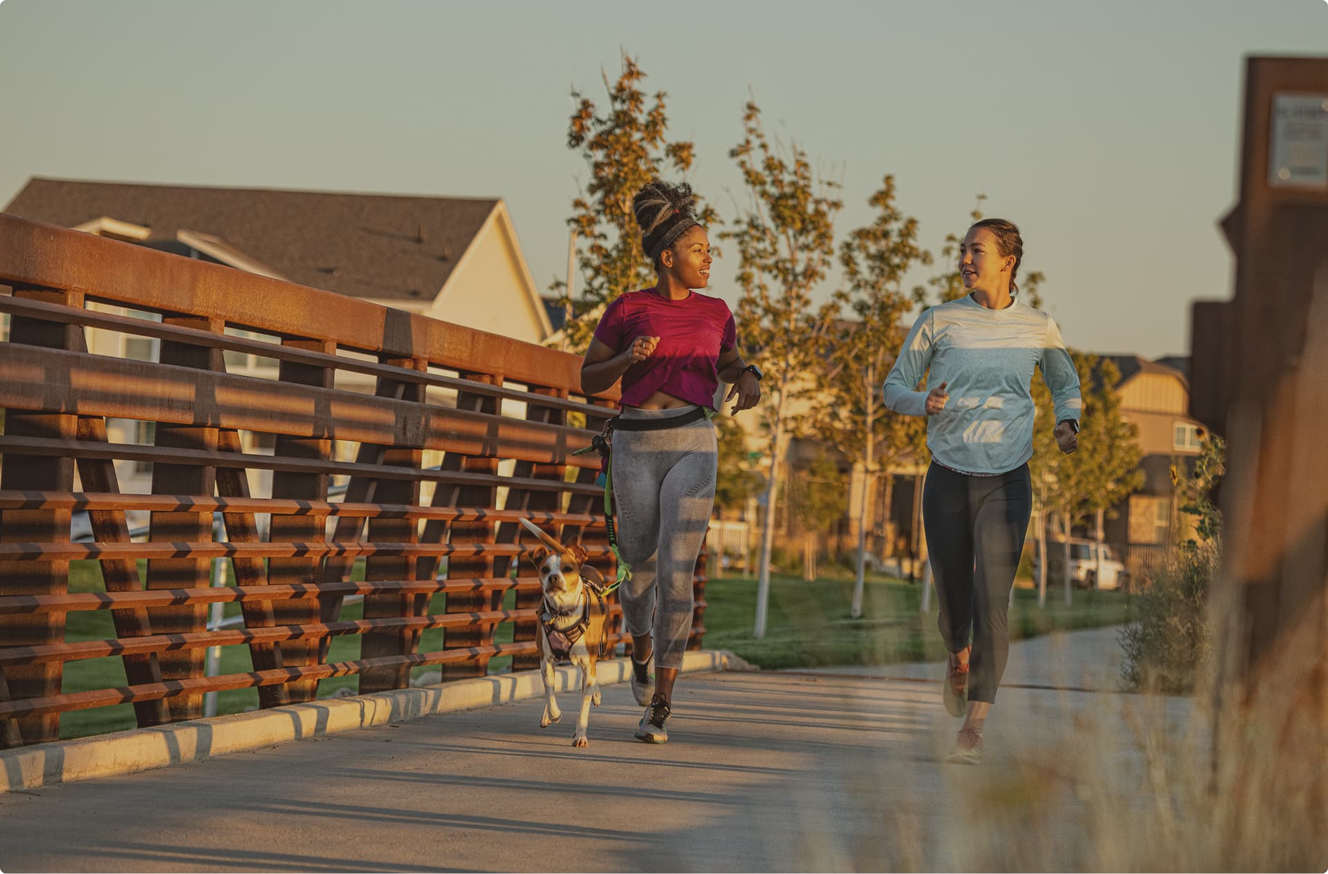 Two women jog on a bridge with a dog on a leash during daylight, passing by trees and new homes in Aurora, CO visible in the background.
