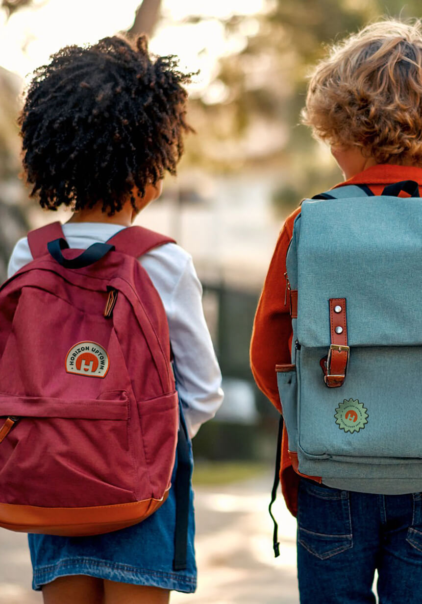 Two children with backpacks walk outdoors in a neighborhood of new construction homes in Aurora CO. One wears red, the other light blue, as they stroll beneath trees and sunlight in this inviting community.