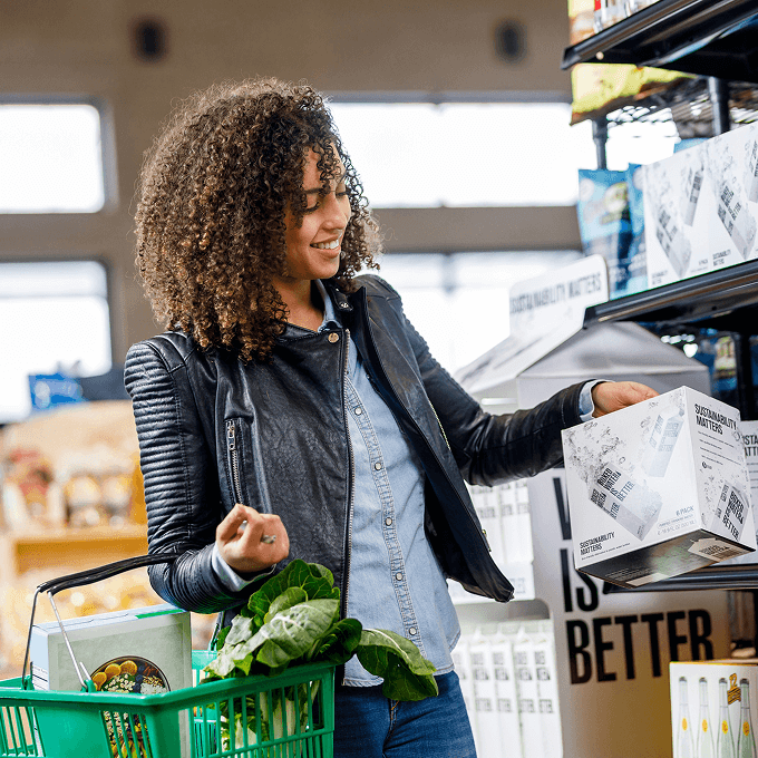 A woman with curly hair holds a green shopping basket of produce and examines a boxed item in a grocery store aisle near Horizon Uptown Aurora, one of the vibrant new home communities in Aurora, CO.