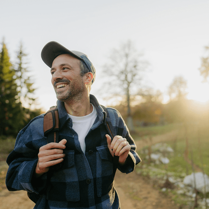 A man wearing a cap and plaid jacket smiles while walking outdoors with a backpack, exploring the scenic trails near new construction homes in Aurora, CO. Trees and sunlight are visible in the background.