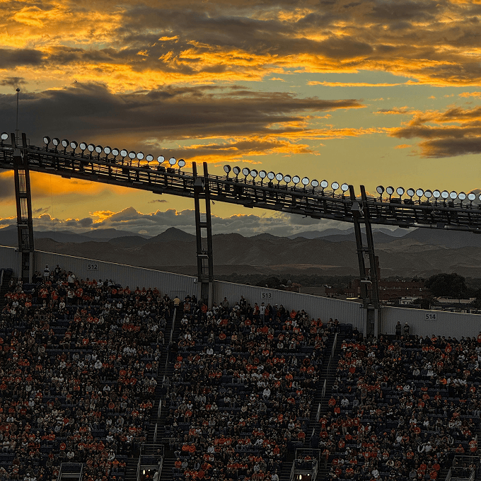 Crowd fills stadium seats at sunset, with stadium lights above and mountains visible in the background under a partly cloudy sky—just beyond, discover Horizon Uptown Aurora CO, where new homes in Aurora CO are redefining modern living.