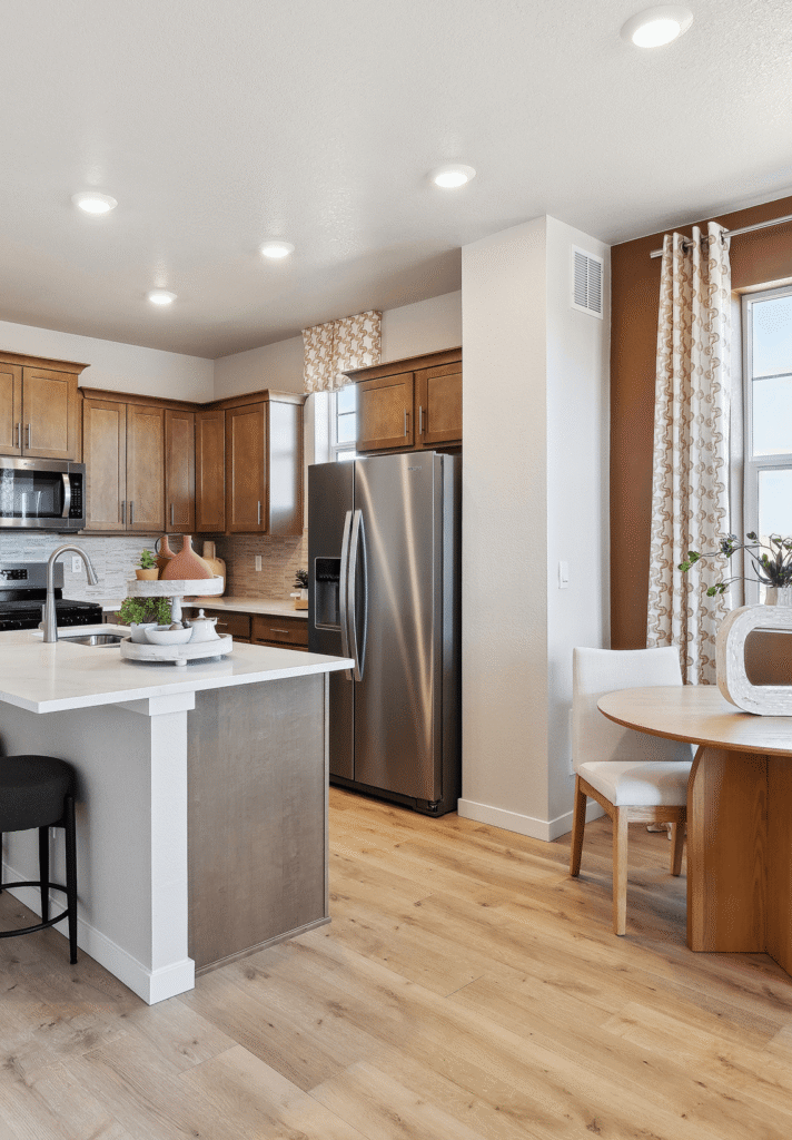 Modern kitchen with wood cabinets, stainless steel appliances, an island with a sink, and a round dining table with chairs on light wood flooring—perfect for those seeking new homes in Aurora CO.