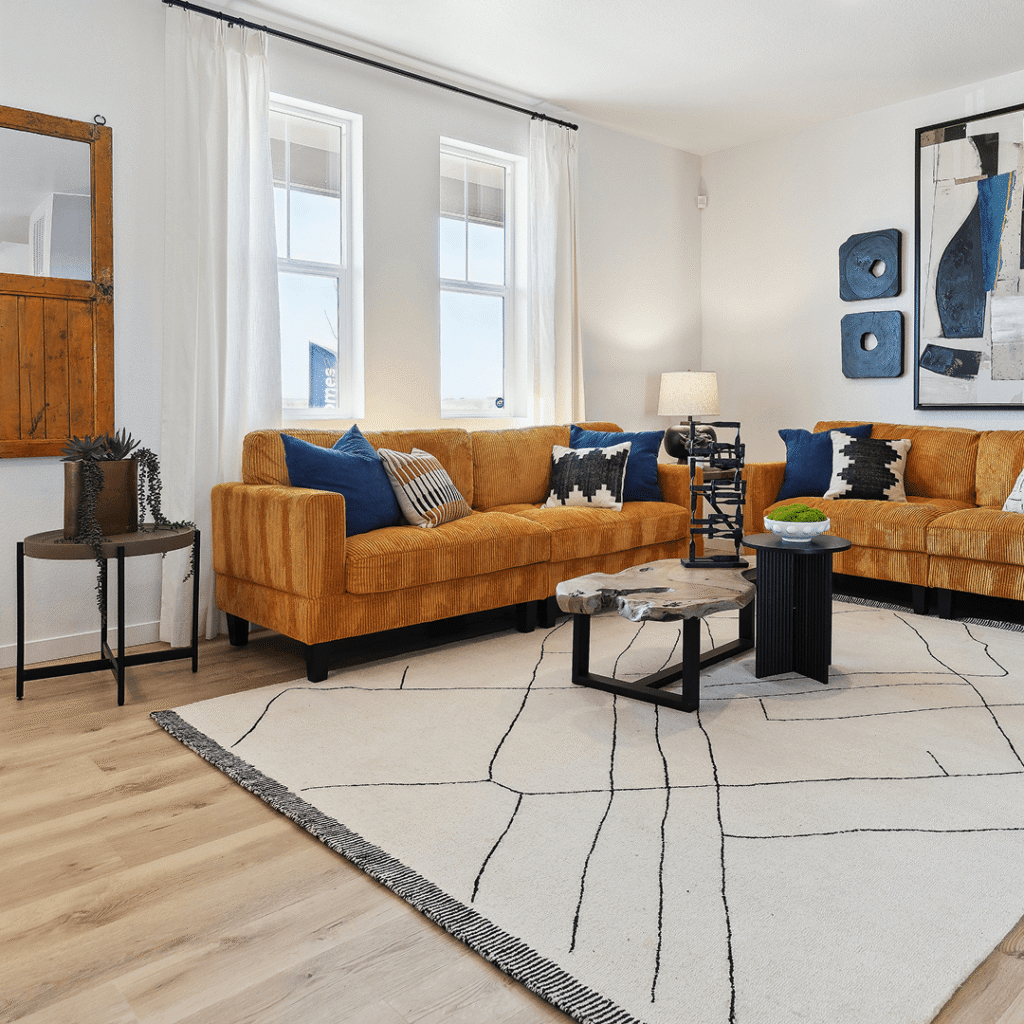 Modern living room in Horizon Uptown Aurora with mustard yellow sectional sofas, blue and black accent pillows, abstract wall art, geometric coffee tables, and a white area rug with black lines.