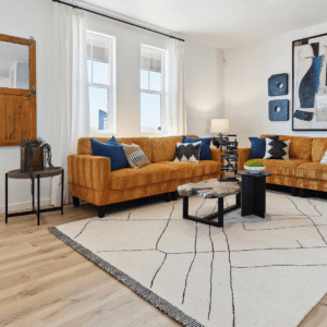 Modern living room in Horizon Uptown Aurora with mustard yellow sectional sofas, blue and black accent pillows, abstract wall art, geometric coffee tables, and a white area rug with black lines.