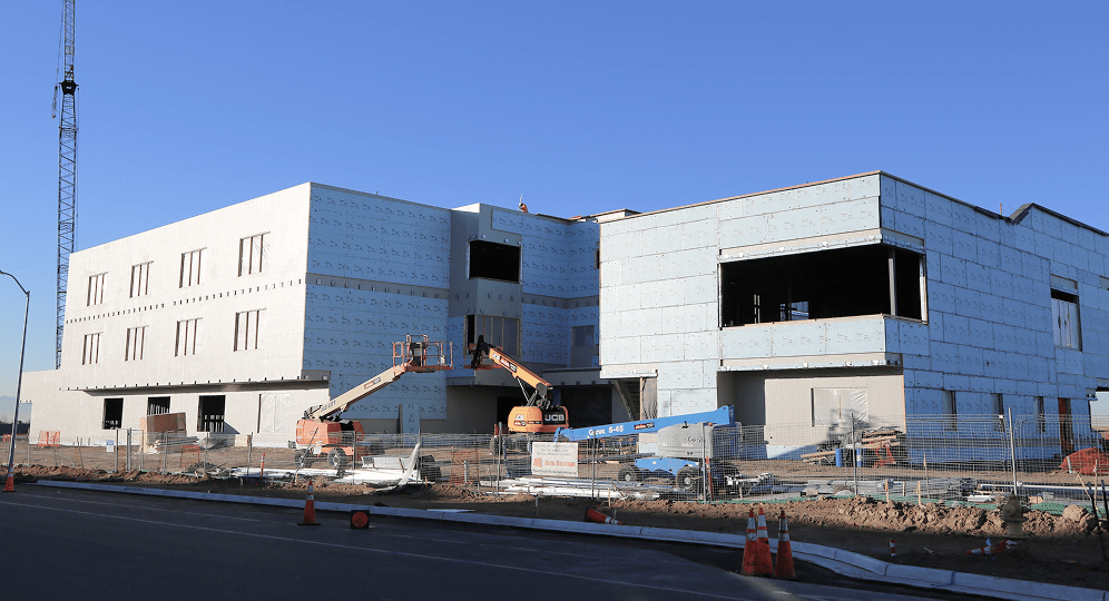 A large, partially constructed building with exposed insulation, construction equipment, and materials scattered around the site under a clear blue sky in Horizon Uptown Aurora, one of the top new home communities in Aurora CO.
