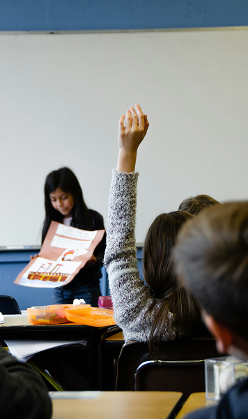 A student stands at the front of a classroom presenting a project, while another student seated raises their hand to participate—a scene reflecting the bright future of learning in Horizon Uptown Aurora CO new home communities.
