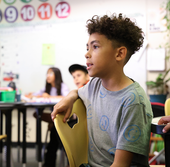 A boy sits attentively in a classroom, with other students and a whiteboard featuring colorful numbered circles in the background—reflecting the vibrant community found near new construction homes in Aurora, CO.
