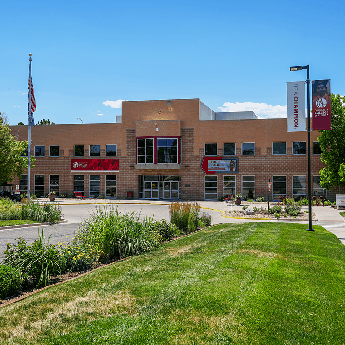 A brick academic building with large windows and banners stands by landscaped greenery and an American flag under a clear blue sky, reflecting the welcoming spirit of Horizon Uptown Aurora CO.