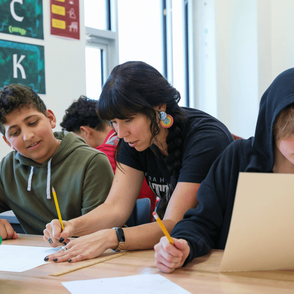 A teacher assists two students with their assignments at a classroom table; all are holding pencils and focusing on their work, much like families exploring new homes in Aurora CO, coming together to build bright futures.
