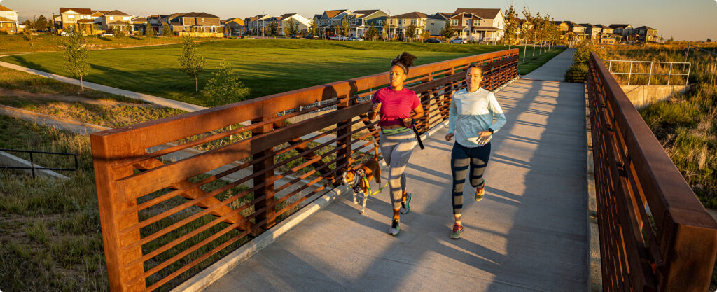Two women jog side by side with a dog on a leash across a pedestrian bridge in a suburban neighborhood, with new construction homes in Aurora CO and a grassy field visible in the background.