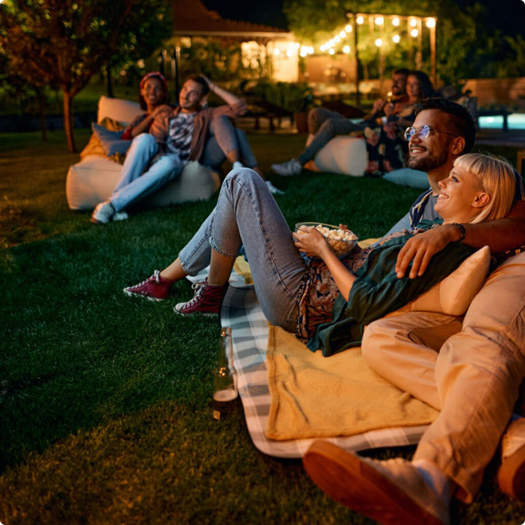 A group of people sit on blankets and beanbags in a backyard at night, watching an outdoor movie and eating popcorn—a perfect evening for those enjoying new homes in Aurora CO, surrounded by string lights and trees.