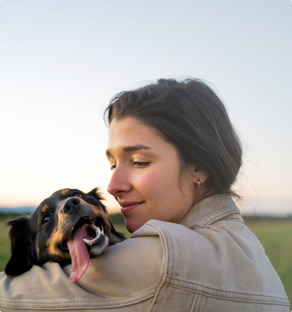 A woman in a beige jacket holds a black and brown dog with its tongue out, outdoors under a clear sky—capturing the joyful spirit of Horizon Uptown Aurora CO and the vibrant lifestyle near new homes in Aurora CO.