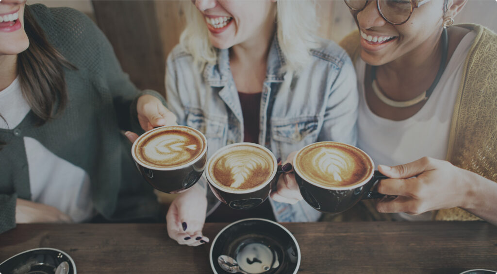 Three people sit together at a table in Horizon Uptown Aurora, each holding a cup of latte with latte art, smiling and appearing to enjoy their time while discussing new home communities nearby.