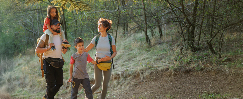 Four people—two adults and two children—walk together on a wooded trail near Horizon Uptown Aurora CO. One child rides on an adult’s shoulders while the others walk side by side, surrounded by trees and grass.