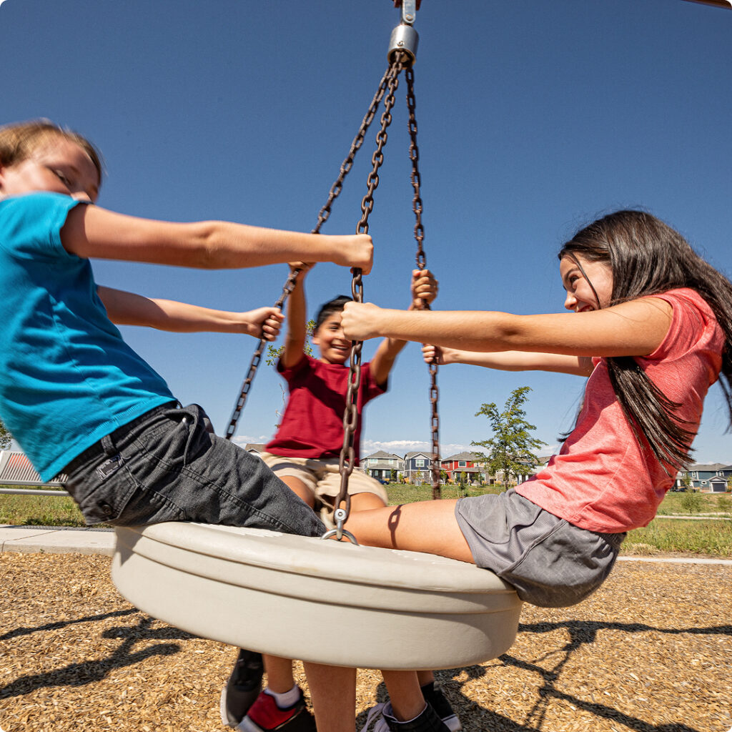 Three children are sitting on a round swing, spinning and laughing under a clear blue sky at Horizon Uptown Aurora, capturing the playful spirit of this vibrant new home community.