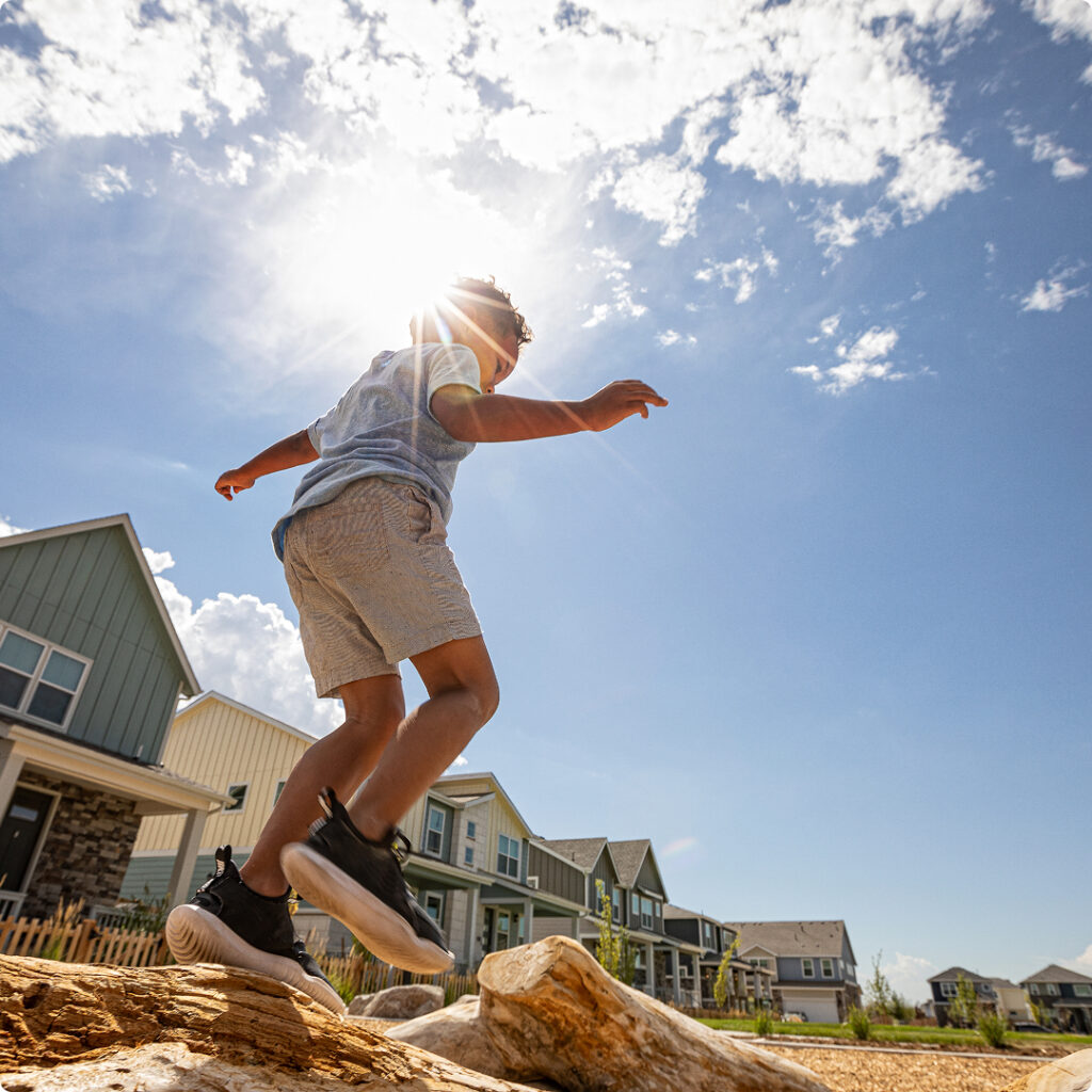 A child balances on a large log in a residential neighborhood with new homes in Aurora, CO, enjoying the sunshine and houses of Horizon Uptown Aurora in the background.