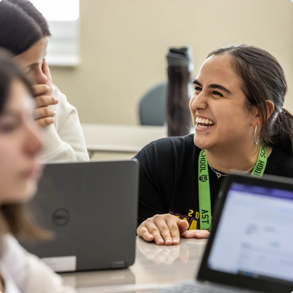 A student wearing a green lanyard smiles and talks with a classmate at a table with laptops in a classroom, discussing their upcoming research project on Horizon Uptown homes for sale.