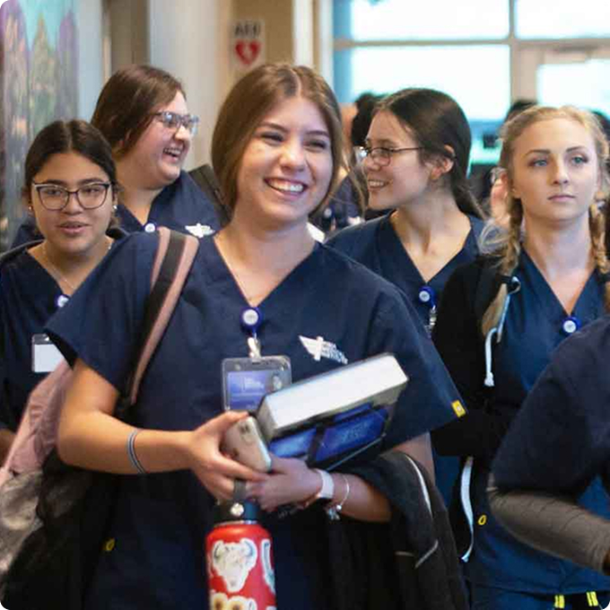 A group of nursing students in navy scrubs walk down a hallway at Horizon Uptown Aurora CO, some smiling and carrying books and water bottles.
