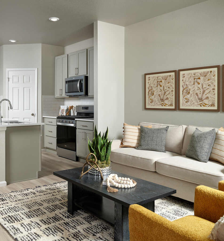Modern living room with a beige sofa, gray and yellow pillows, black coffee table, patterned rug, indoor plant, and a view of a kitchen with white cabinets and stainless steel appliances.